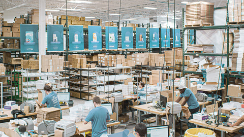 A large warehouse with shelves filled with boxes and electronic devices. Several workers in blue shirts are sitting at tables, operating computers and packing items. Banners displaying product images hang from the ceiling.