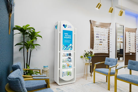 Optometrist office interior with a display stand labeled Daily Eye Care Simplified featuring eye care products. Blue chairs and decorative plants are arranged in the room, with a wall displaying various eyeglasses in the background.
