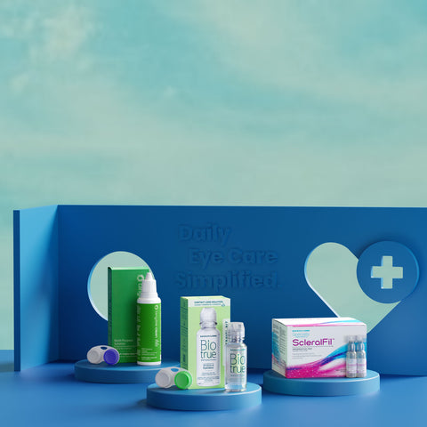 A display features two contact lens cases, contact solution bottles labeled BioTrue, and a ScleralFil box. The backdrop is blue with a heart and plus sign design, featuring the text Daily Eye Care Simplified embossed on it.