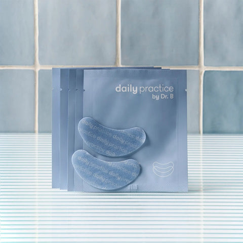 Four light blue packets of Daily Practice Brighten & Hydrate Eye Patches, labeled daily practice by Dr. B, are displayed with eye masks in front. They rest on a blue and white striped surface, backed by a light blue tiled wall, promising to instantly refresh and reduce puffiness.