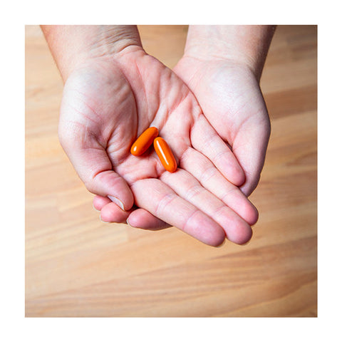 Hands holding two orange oblong pills over a wooden floor background, offering dry eye relief with EyePromise® EZ Tears, made with Omega-3s, Turmeric, and Green Tea Extract.