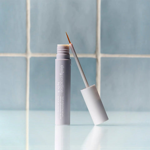 A tube of Daily Practice Lash Enhancing Serum for fuller, longer, healthier lashes (4ml) sits open on a reflective surface with a thin applicator leaning against it. Light blue tiled walls highlight its cutting-edge formula infused with biomimetic peptides for optimal lash growth.