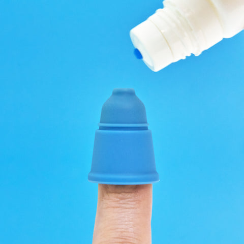 A finger, capped with a blue silicone piece resembling an ice cream cone, captures the whimsical essence of a Magic Touch Eye Drop Applicator. In the top right corner, a bottle featuring a blue nozzle cap is set against a bright blue backdrop.
