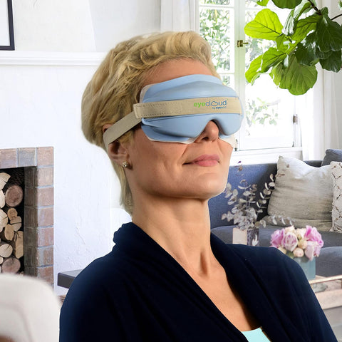 A woman relaxes in a bright living room wearing the EyeEco EyeCloud Home Treatment Kit (Blue) eye mask, enjoying moist heat therapy for dry eyes with over 600 gel pack uses.
