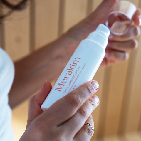 Close-up of a person holding a white bottle labeled Merakim Facial Sunscreen SPF 50 in orange text, designed for sensitive skin. They are removing a clear cap from the pump dispenser. The softly blurred background showcases vertical wooden paneling, hinting at its broad-spectrum protection with zinc oxide.