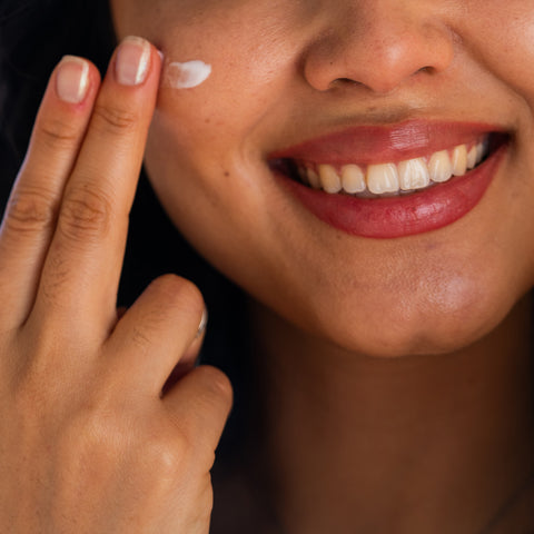 A smiling person applies Merakim Facial Sunscreen SPF 50 to their cheek with fingers, revealing white teeth and red lipstick against a blurred background, while enjoying the protective benefits on sensitive skin.
