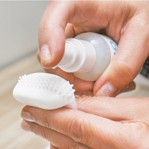 A close-up shows hands applying MyboClean Lids and Lash Gel onto a MYBOClean eyelid cleansing brush. The small white silicone brush, ideal for eyelid cleaning, has soft bristles. The persons nails sparkle with glittery polish.