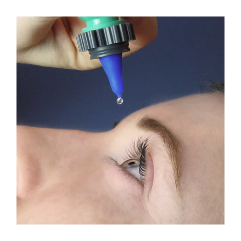 Against a dark blue background, a light-skinned person tilts their head back, as a hand holds the Nanodropper Eye Drop Extender Adaptor poised above their eye, reducing drop size by 60% for less waste and easier application.