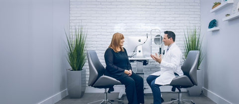 A woman and a man in a white coat sit facing each other in a modern office with a white brick wall. They are engaged in conversation next to a medical device. Tall green plants flank the walls, and shelves with decorations are on the right.