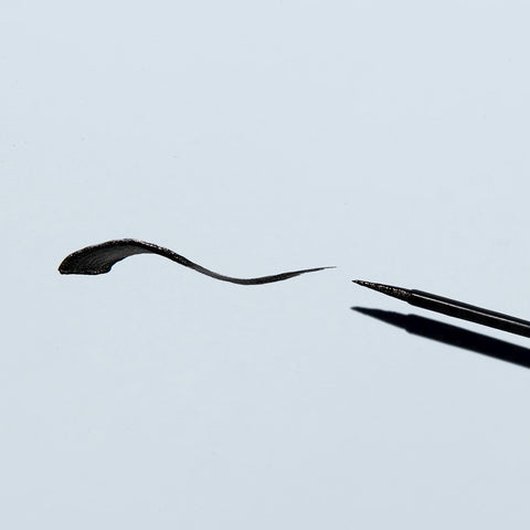 Close-up of the Twenty Twenty Double Duty Precision Eyeliner (Pitch Black) next to a sleek, curved line on a light gray background. The fine bristles offer a precise finish for fuller-looking lashes with each application.