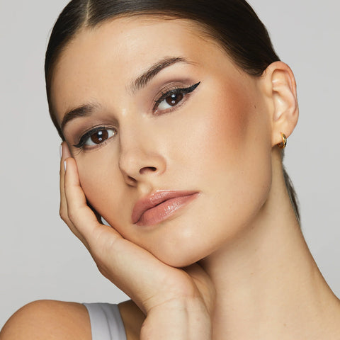 A woman with long dark hair in a sleeveless top poses with her hand on her cheek. Her eyes stand out with Twenty Twenty Double Duty Precision Eyeliner (Pitch Black), fuller lashes, neutral eye makeup, and glossy lips against a soft gray backdrop.