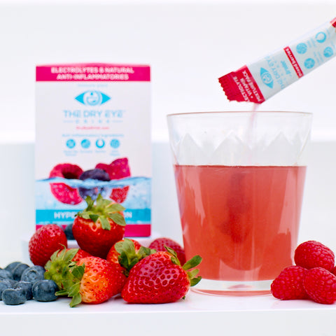 In front of a box of Dry Eye Drink™ Daytime by Bruder, a glass of pink beverage with mixed berries powder pouring into it sits among fresh strawberries, blueberries, and raspberries. Infused with anti-inflammatory ingredients, it provides doctor-formulated electrolyte hydration for eye health.