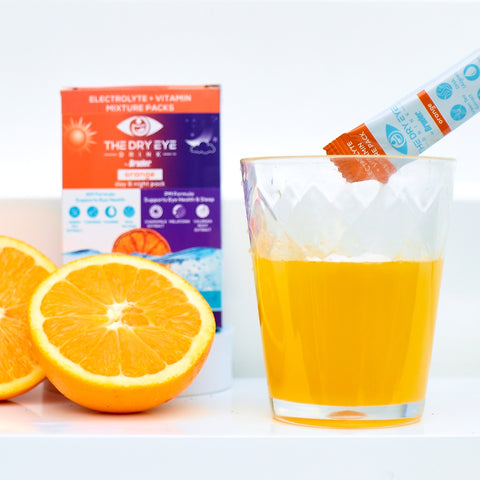 A clear glass with an orange drink showcases a packet of Dry Eye Drink™ Complete Set by Bruder being poured, promising dry eye relief. Two orange halves and the product box highlight its electrolyte and vitamin mix for optimal eye health. Available in 20 packets, 10 each for day and night hydration.