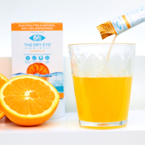 A glass of orange drink from a sachet is being poured, promising eye health and refreshing taste. Sliced oranges are beside it. In the background, a box labeled Bruder Dry Eye Drink™ Daytime shows an orange splash, emphasizing its doctor-formulated hydrating formula for dry eye relief.