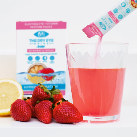 A glass of pink Dry Eye Drink™ Daytime, a strawberry lemonade powdered supplement by Bruder for eye health, is being prepared. Fresh strawberries and a lemon half are nearby, with the product box in the background featuring fruit images and text.