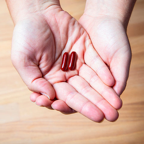Hands holding EyePromise Macular Health Complete AREDS 2 capsules to enhance vision and support macular health, over a wooden floor background.