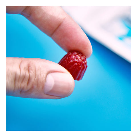 A hand holds a small, wild berry FitEyez gummy against a bright blue background, pinched between the thumb and index finger.