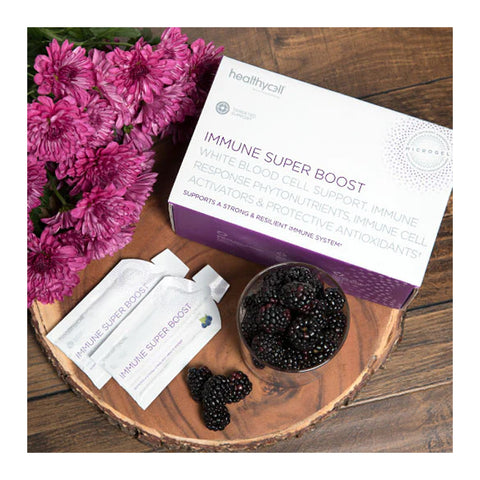 A HealthyCell Immune Support Boost Gel Packs box is on a wooden surface with two sachets nearby. A bowl of blackberries and loose ones accompany it, while purple flowers and elderberry blossoms are arranged at the top left corner.