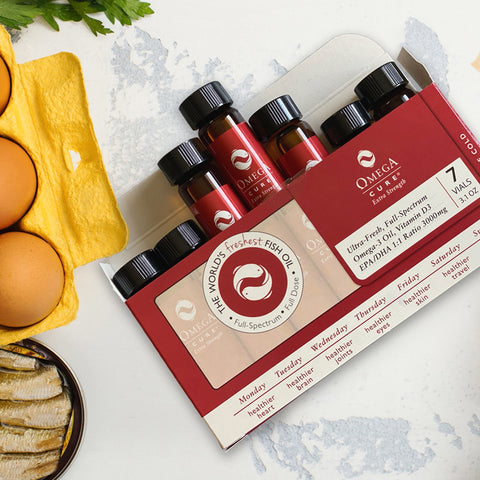 A display of Omega Cure Extra Strength Fresh Liquid Omega from Omega 3 Innovations, in a weeks supply package with cold shipping, sits beside brown eggs, sardines, and greenery on a marble surface. The packaging proudly states The Worlds Freshest Fish Oil.