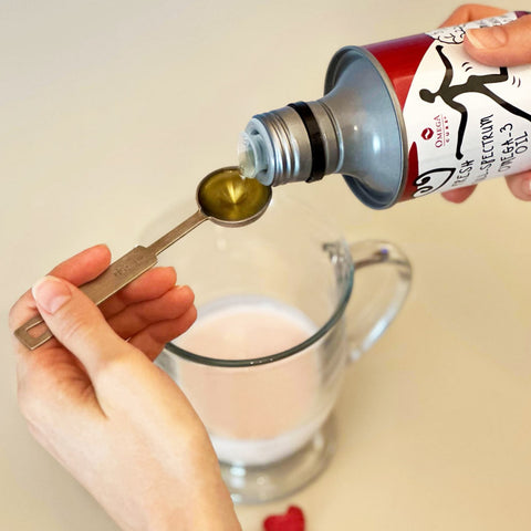 Someone pours Omega Cure Omega 3 Fish Oil from a bottle into a teaspoon over a clear glass of light pink liquid. The Omega 3 Innovations label ensures an omega-3 dose with no fishy taste, set against a light beige backdrop.