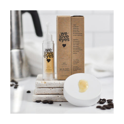 The We Love Eyes - AM Eyelid Gel by We Love Eyes, accompanied by a dropper, rests on white stone slabs. It promises relief for MGD and dry eyes. Nearby is its box. Scattered coffee beans and a cream-toned lid appear in the foreground, with a gleaming sink faucet as the backdrop.