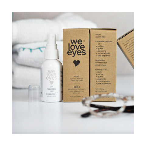 A We Love Eyes Calm Eyelid & Eyelash Cleansing Spray with HypoChlorous (50mL) bottle and box are on a white surface, showcasing its vegan, cruelty-free formula. Chamomile adds soothing properties, while blurred glasses and towels in the background evoke everyday elegance.
