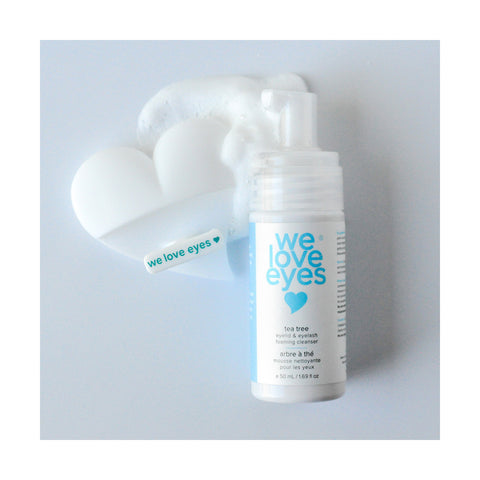 A bottle of We Love Eyes tea tree eyelid and eyelash foaming cleanser stands in heart-shaped white foam. A tag reads We Love Eyes on its left. Ideal for eye care, it pairs perfectly with the We Love Eyes Eyelid Scrub Mitt against a light gray backdrop.