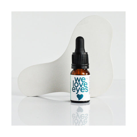 A small amber dropper bottle of We Love Eyes Lashfull Thinking Lash + Brow Follicle Oil 10mL sits on a reflective white surface, with a blue heart label, set against an abstract white backdrop to emphasize its benefits for lashes and brows.