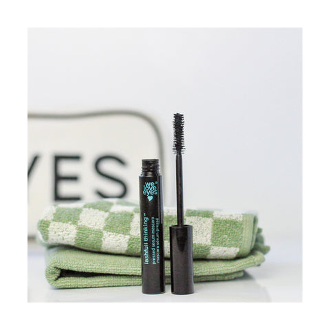 A tube of We Love Eyes Lashfull Thinking BLACK Pressed Serum Mascara with Widelash stands open in front of two green and white checkered towels, while a sign reading EYES is partially visible in the background. The formula enhances eyelid skin health for a refreshed look.