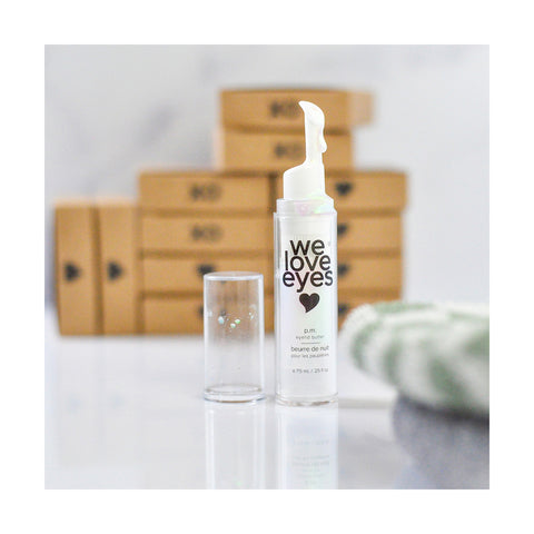 A pump bottle of We Love Eyes - P.M. Eyelid Butter (7.5ml) sits elegantly with its cap nearby, promising hydration for dry eyes. The background features blurred brown boxes and a hint of green and white cloth in the corner.