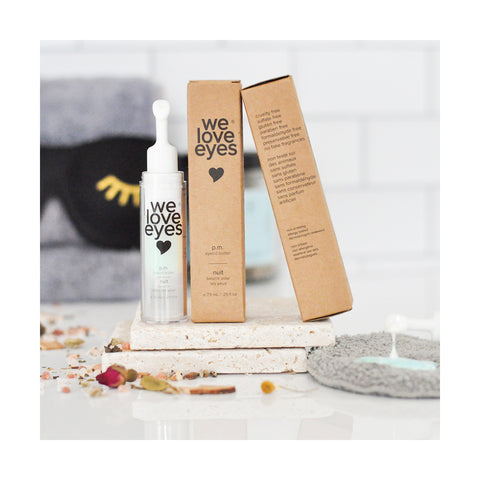 A dropper bottle labeled We Love Eyes - P.M. Eyelid Butter - 7.5ml sits on stone tiles with its brown packaging. Designed for dry eyes and fine lines, an eye mask and dried petals enhance the serene setting.
