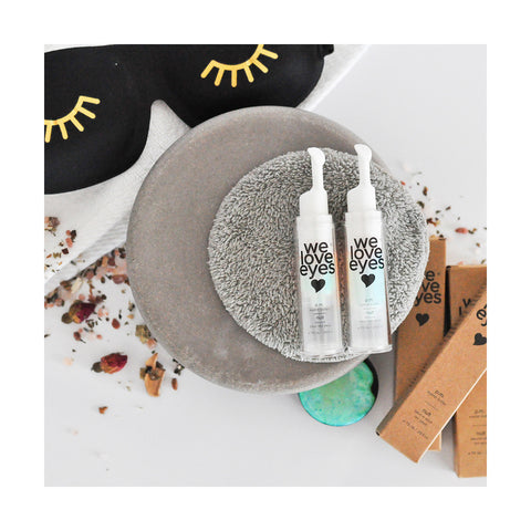 Flat lay of We Love Eyes - P.M. Eyelid Butter alongside another white dispenser on a gray towel, with brown boxes, a black eyelash sleep mask, and dried petals scattered around, creating a soothing atmosphere for tackling dry eyes and fine lines.