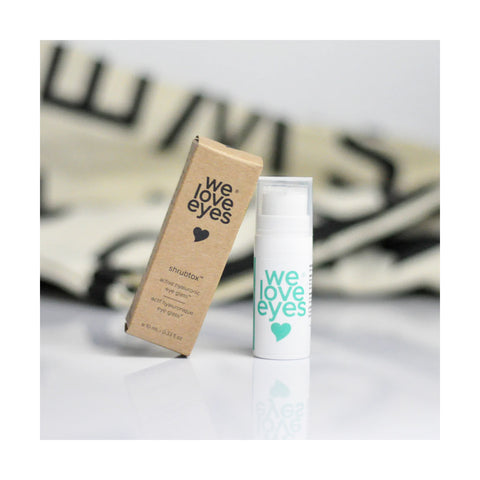 A We Love Eyes cardboard box beside a white bottle labeled Shrubtox Active Hyaluronic Eye Glass (10mL, pump) promises an eyelid skin barrier boost. The softly blurred background highlights them as essential allies against fine lines and wrinkles.