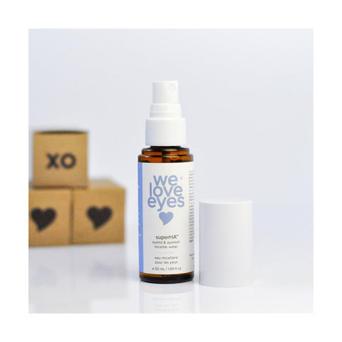 A 50mL spray bottle of We Love Eyes superHA Eyelid & Eyelash Micellar Water Make-up Remover with a white cap sits beside three wooden blocks—one marked XO and another with a heart symbol—on a white surface. Infused with tea tree water, its ideal for eyelid skin health and gentle makeup removal.
