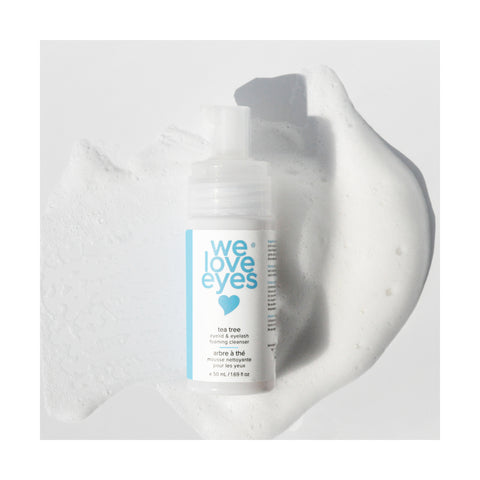 A 25mL We Love Eyes Tea Tree Eyelid & Eyelash Foaming Cleanser bottle sits on a textured white foam background. The label features tea tree foaming cleanser in blue and gray text, emphasizing its refreshing tea tree oil formula.