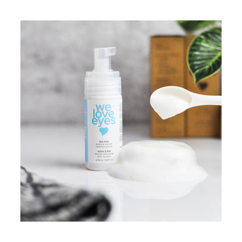 The We Love Eyes Tea Tree Eyelid & Eyelash Foaming Cleanser 25mL Mini Size sits on a white surface beside a plastic spoon. In front, foam is piled, while a green plant with blurred leaves adorns the background.