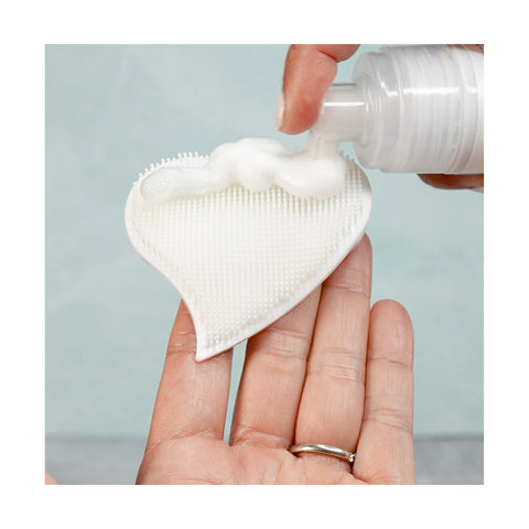 A hand applies We Love Eyes Tea Tree Eyelid & Eyelash Foaming Cleanser onto a heart-shaped silicone scrubber with delicate bristles, ensuring a gentle cleanse against a soft mint green backdrop.