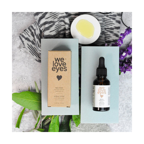 A flat lay showcases We Love Eyes - All Natural Tea Tree Eyelid Cleansing Oil in a dark glass dropper bottle with its brown box on gray surfaces. Accompanied by green leaves, a purple flower, jojoba oil dish, and fabric, its ideal for nourishing Meibomian oil glands.