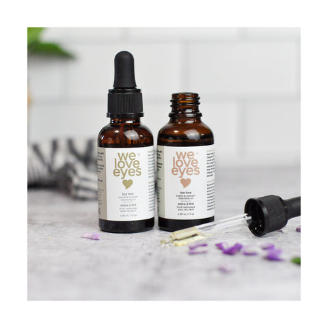 Two amber bottles, labeled We Love Eyes- All Natural Tea Tree Eyelid Cleansing Oil, sit on marble. One is open with its dropper beside it, infused with Tea Tree Oil. Purple petals and a blurred plant create a tranquil backdrop.