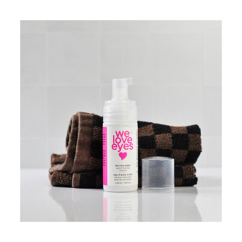 The We Love Eyes Tea Tree Water Eyelid Foaming Cleanser by We Love Eyes, with its clear cap, rests in front of a folded brown checkered towel on a reflective surface against a tiled wall. This pH-balanced cleanser embodies refreshing tranquility.