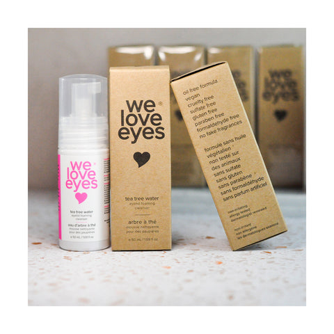 A bottle of We Love Eyes Tea Tree Water Eyelid Foaming Cleanser stands beside its cardboard packaging, featuring a pink heart logo. Ingredients and benefits are listed on the box, with two other packages subtly blurred in the background.