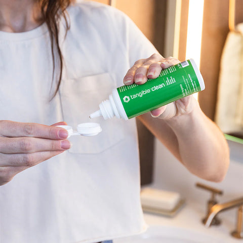Person dispensing solution from a green bottle labeled tangible clean onto a contact lens case lid. They are wearing a white shirt in a bathroom setting with a mirror and towel in the background.