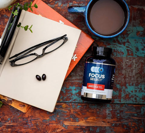 A pair of glasses rests on an open notebook with two black pills. Nearby are a blue mug filled with a dark beverage and a bottle labeled Focus Select AREDS2. The background features a rustic wooden surface.