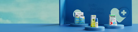 Three eye care products are displayed on blue pedestals. The background features a stylized eye and cross symbol with the text Daily Eye Care Simplified. The setting is minimalistic with a light blue backdrop.