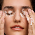 A close-up shows a person with closed eyes, gently massaging Daily Practices Purifying Foam Cleanser into their skin. Their fingers are coated in white foam, and they appear relaxed. The background is not visible.