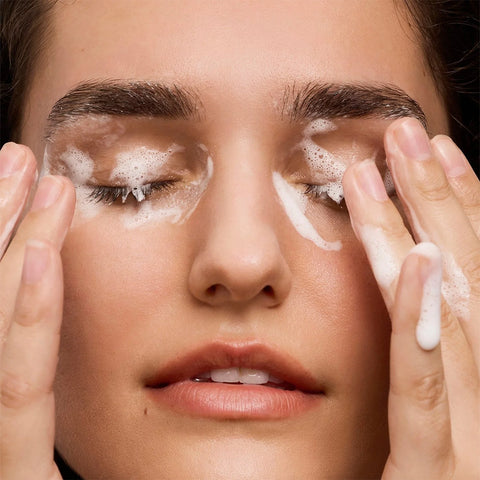 A close-up shows a person with closed eyes, gently massaging Daily Practices Purifying Foam Cleanser into their skin. Their fingers are coated in white foam, and they appear relaxed. The background is not visible.