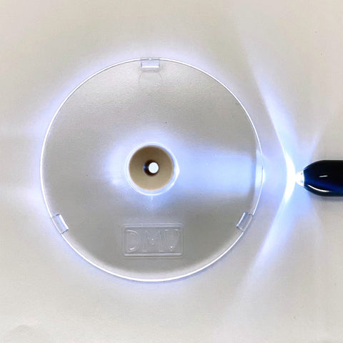 A DMV Scleral Contact Lens Inserter, resembling a clear plastic CD with a central hole, is lit by a small blue flashlight on the right. The letters DMV are embossed on it, and it rests elegantly on a white surface.