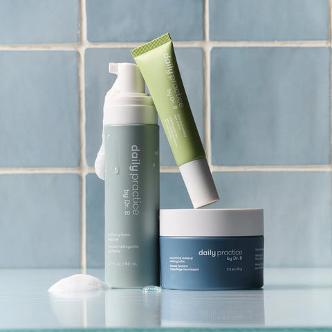 Against a tiled background are three skincare products: a green purifying foam bottle, an exfoliating face mask tube, and a blue nourishing moisture cream jar. Enhance your routine with Daily Practices anti-aging eye cream from the Three Step Eye Care Routine set.