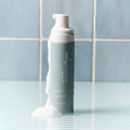 A gray bottle of Daily Practice purifying foam cleanser, part of the Three Step Eye Care Routine Bundle, stands on a striped surface with foam spilling over. Enhance your eye care on light blue tiles with this refreshing cleanser.