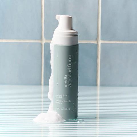 A gray bottle of Daily Practice purifying foam cleanser, part of the Three Step Eye Care Routine Bundle, stands on a striped surface with foam spilling over. Enhance your eye care on light blue tiles with this refreshing cleanser.